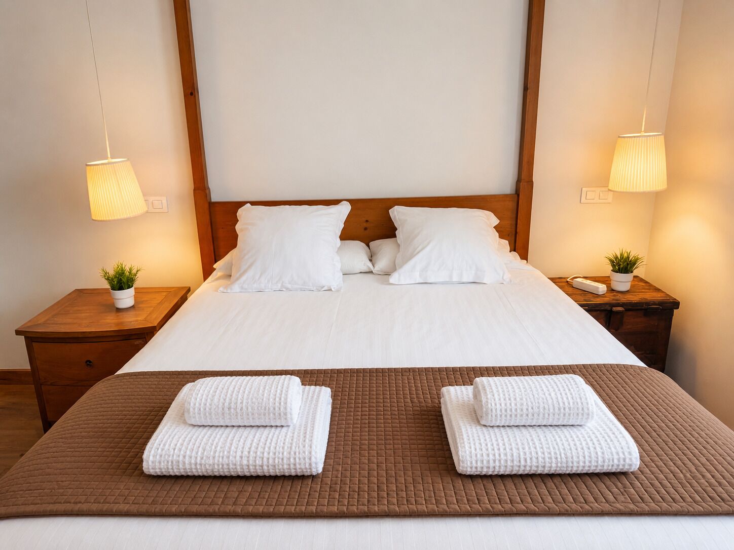 A neatly made bed with white linens, two pillows, a brown quilted bedspread, and two folded towels on a wooden nightstand with a small potted plant.
