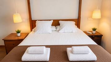 A neatly made bed with white linens, two pillows, a brown quilted bedspread, and two folded towels on a wooden nightstand with a small potted plant.