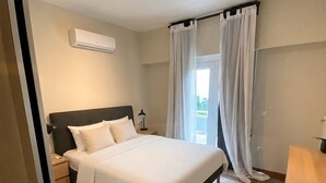 A bedroom with a bed, bedside tables, an air conditioning unit, and a window with curtains. - Solea by Neuvel (Glyfada)