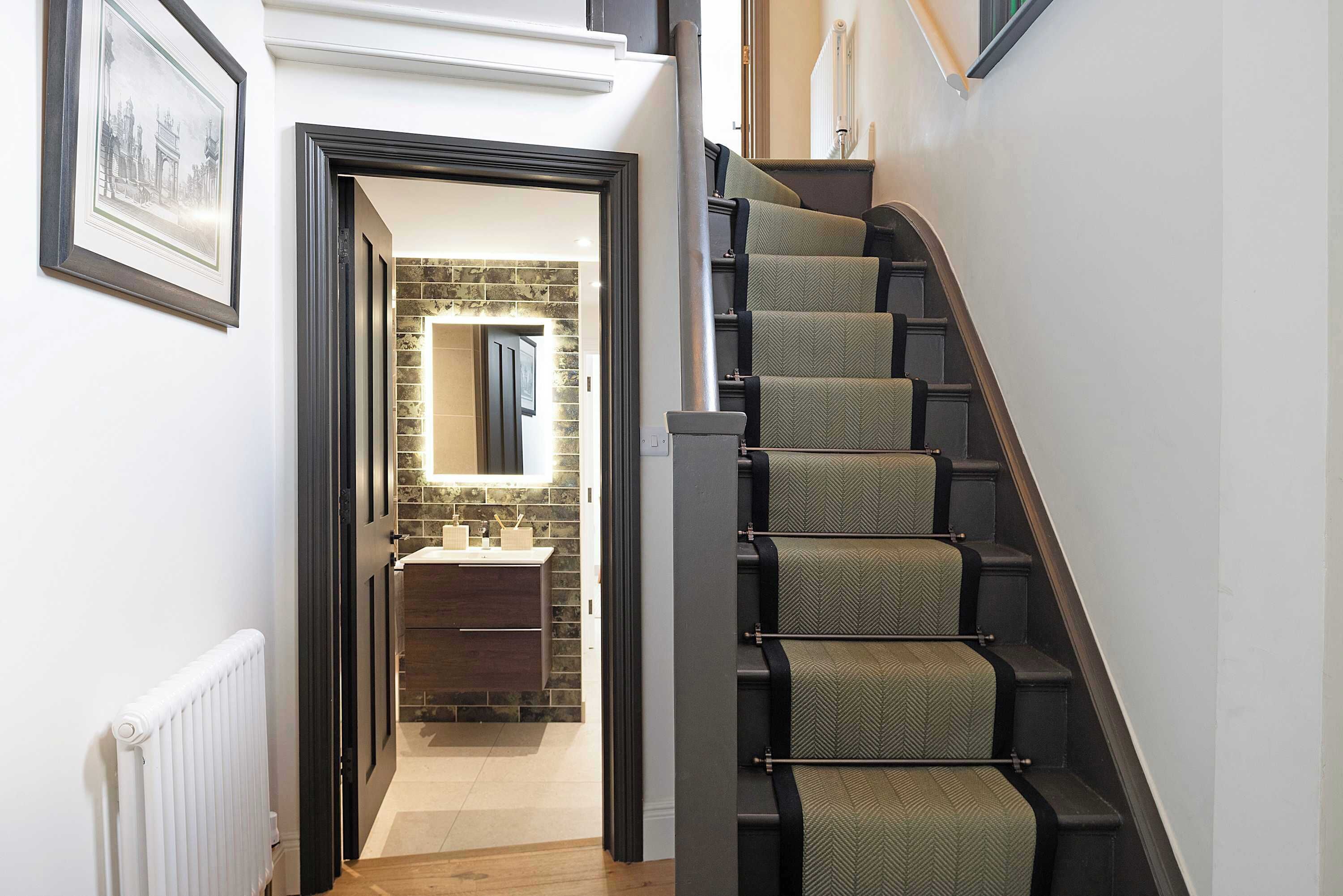 Un escalier doté d’une rampe et de marches recouvertes de moquette, menant à une salle de bain dont un mur est recouvert de carreaux et qui contient un miroir.