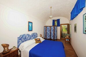 A bedroom with a bed, a nightstand, a lamp, a window with curtains, and a door leading outside. - 8th-century dwelling near Positano (Nocelle)