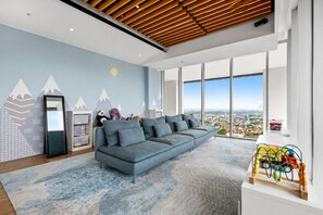 A modern living room with a large gray sofa, a wooden ceiling, and a city view through large windows. - Jasen by Kozystay - Alam Sutera (Tangerang)