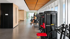 A gym with exercise equipment, a person working out, and a view of the city through large windows. - Jasen by Kozystay - Alam Sutera (Tangerang)