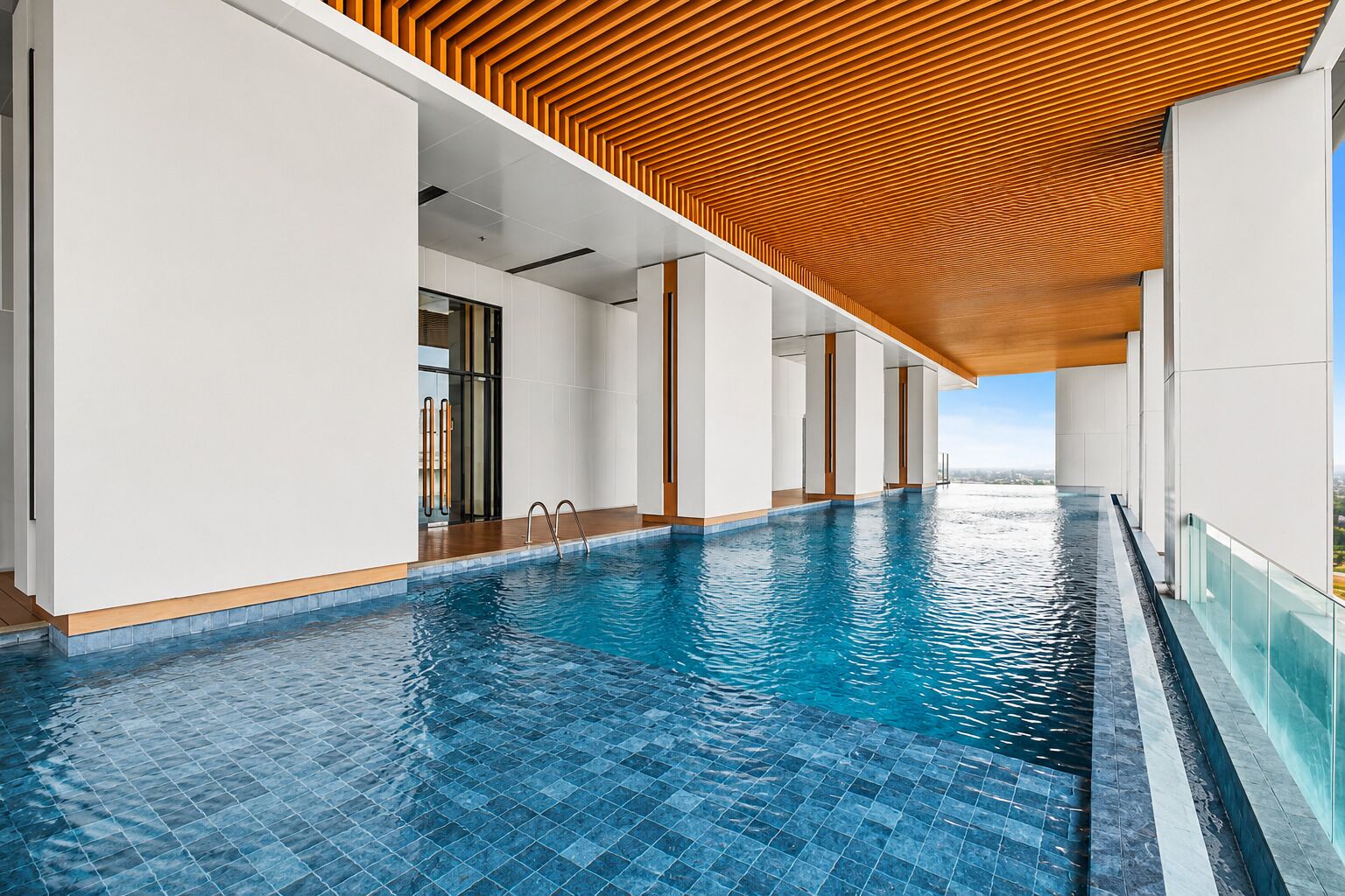 A modern indoor swimming pool with clear blue water, wooden ceiling panels, and a view of the landscape.
