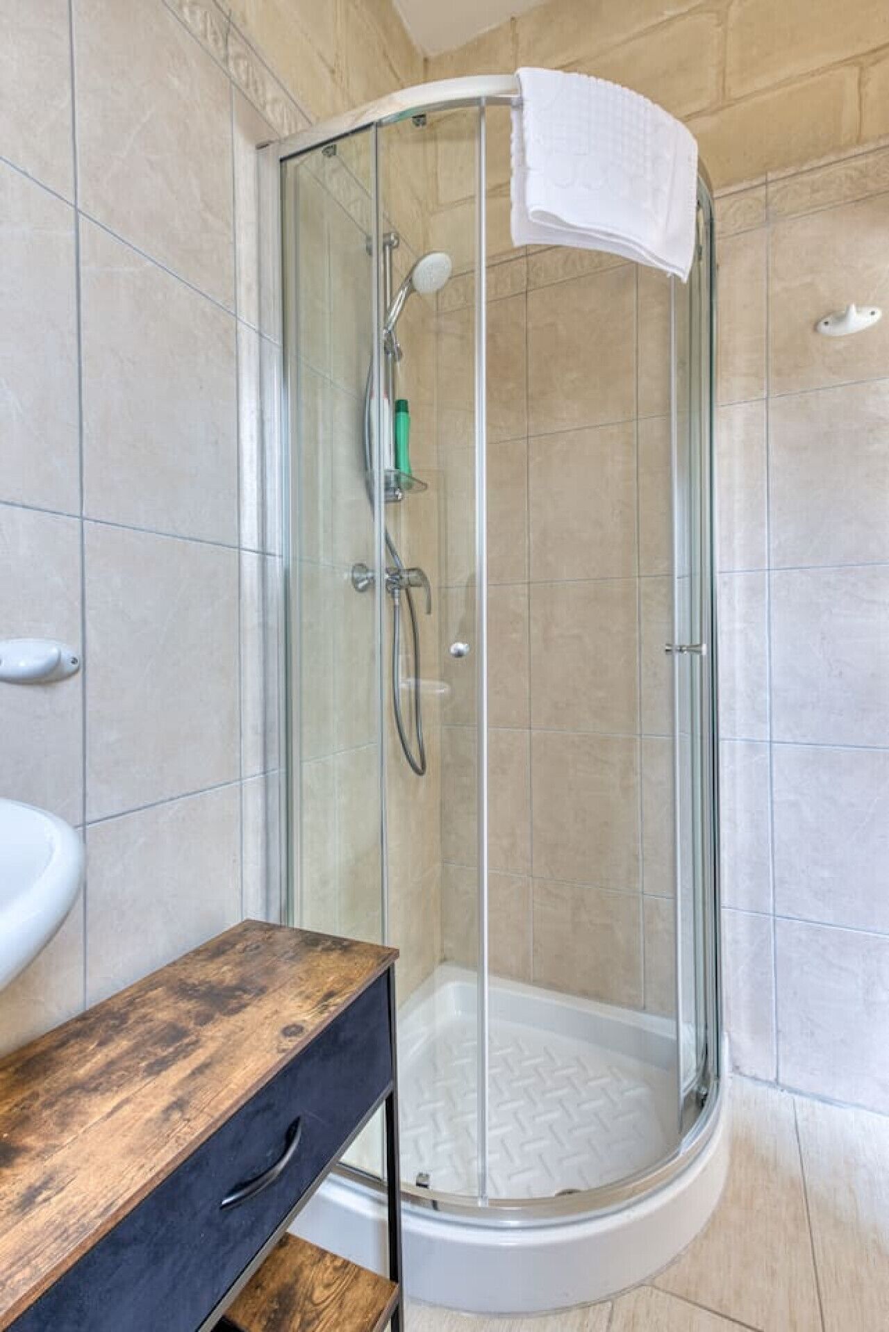 Une douche fermée par des parois de verre, avec une serviette blanche accrochée à la porte, un meuble-lavabo en bois et des murs carrelés.