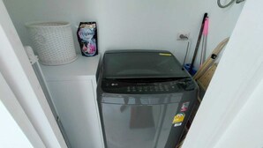 A compact washing machine in a small laundry area with a basket and a hygiene product on the wall. - Kamala Jungle Suite 80m² Balcony, 2 Pools, Gym & Sauna Fast Fiber WiFi (Kamala, Kathu)