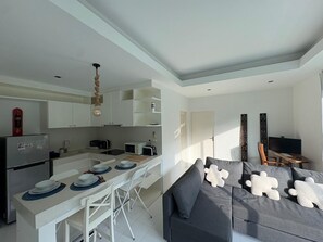 A modern kitchen with white cabinets, a dining area with a table set for two, and a grey sofa with decorative pillows. - Kamala Jungle Suite 80m² Balcony, 2 Pools, Gym & Sauna Fast Fiber WiFi (Kamala, Kathu)
