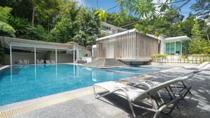 A swimming pool with lounge chairs, a covered seating area, and a modern building with a sloped roof. - Kamala Jungle Suite 80m² Balcony, 2 Pools, Gym & Sauna Fast Fiber WiFi (Kamala, Kathu)