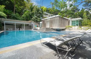 A swimming pool with lounge chairs, a covered seating area, and a modern building with a sloped roof. - Kamala Jungle Suite 80m² Balcony, 2 Pools, Gym & Sauna Fast Fiber WiFi (Kamala, Kathu)
