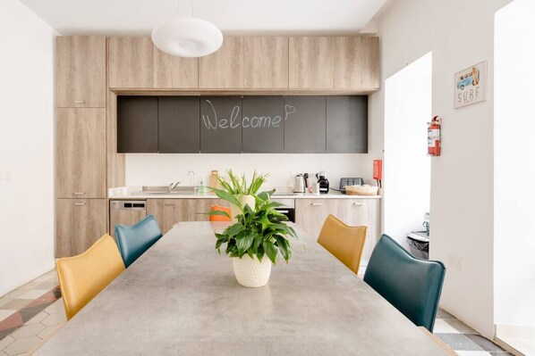 A modern kitchen with a chalkboard saying 'Welcome', a dining table with chairs, and a potted plant.