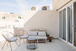 A balcony with a sofa, two chairs, and a small table. There are two air conditioning units on the wall.