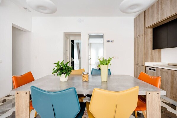 A dining area with a round table, chairs, potted plants, and a kitchen area with a TV.