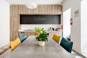 A modern kitchen with a chalkboard displaying 'Welcome', a dining table with chairs, and a potted plant. - Birgu Room -Hammock Vibes Towns House (Tas-Sliema)