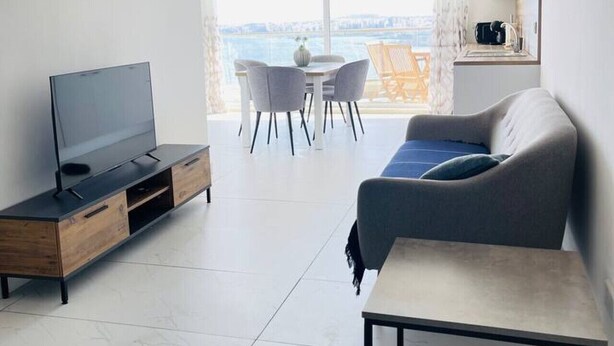 A modern living room with a flat-screen TV, a sofa, a coffee table, and a dining area with chairs. - Blu Mar Sea View Appartment 4 (San Pawl il-Baħar)