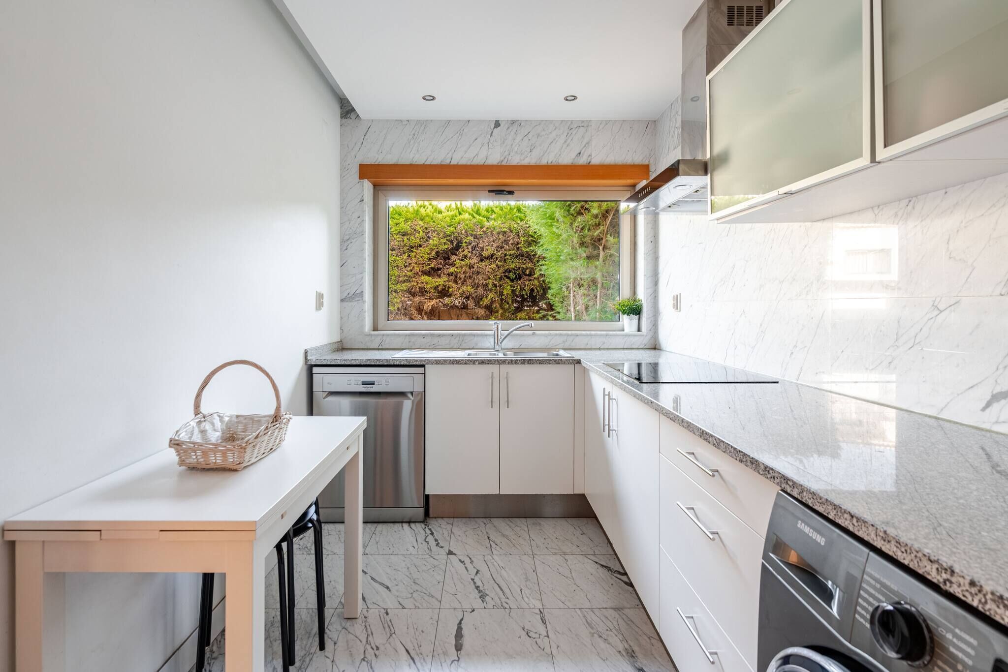 A modern kitchen with a built-in dishwasher, a window with a view of greenery, and a countertop with a Samsung appliance.