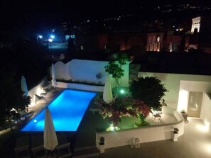 A nighttime view of a swimming pool area with a curved wall, a few lounge chairs, and potted plants. - Villa LuxL Family House (Mesaria)