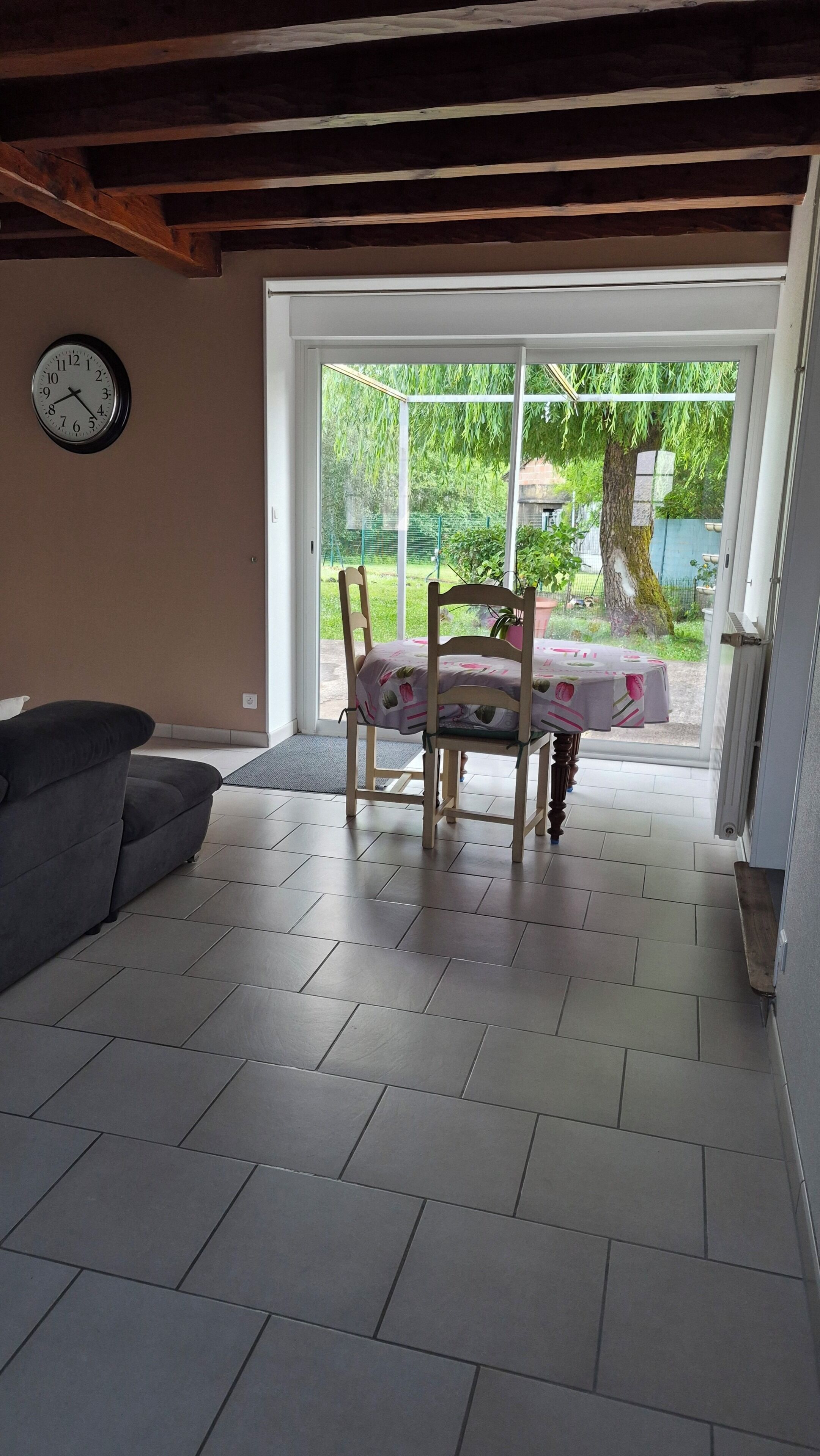 Een kamer met een eettafel en stoelen, een bank, een klok aan de muur en uitzicht op het buitenlands landschap door een glazen deur.