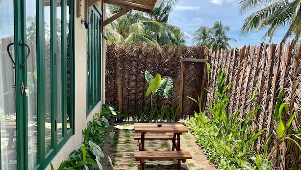A covered patio with a wooden table and benches, surrounded by lush greenery and a wooden fence.