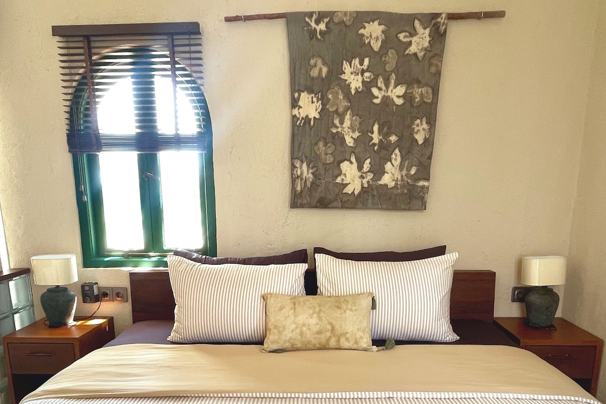 A hotel room with a large bed, two bedside tables, a window with blinds, and a wall-mounted artwork.