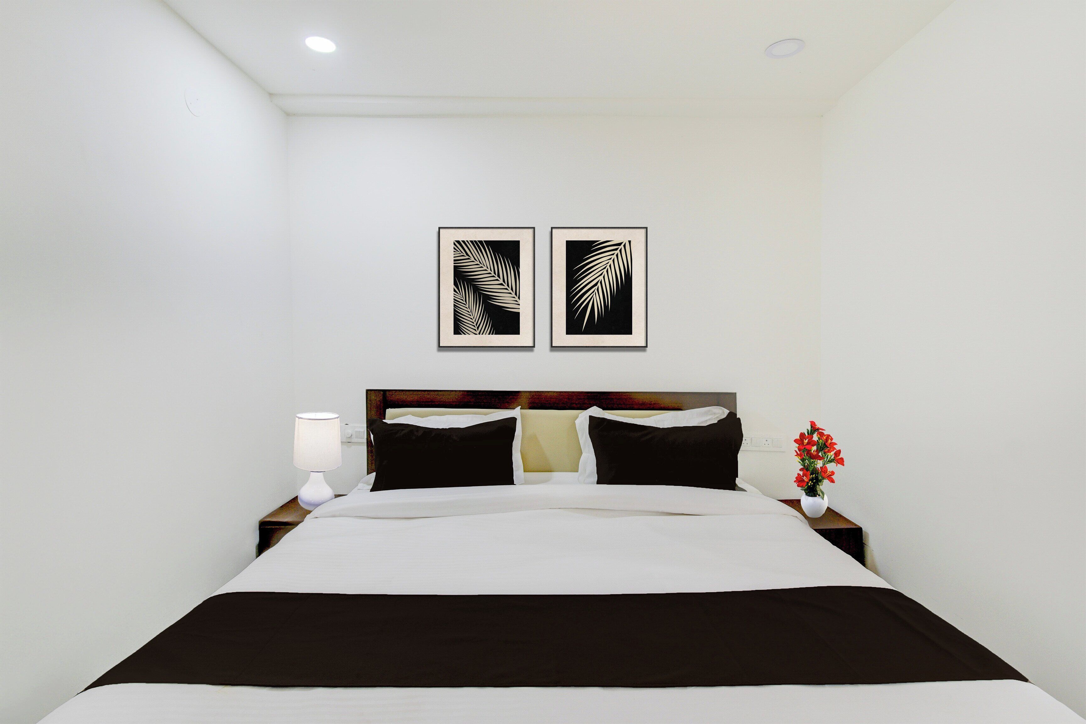 Quarto de hotel com cama, mesa de cabeceira com abajur, duas obras de arte com folhas na parede e um vaso com flores na mesa de cabeceira.