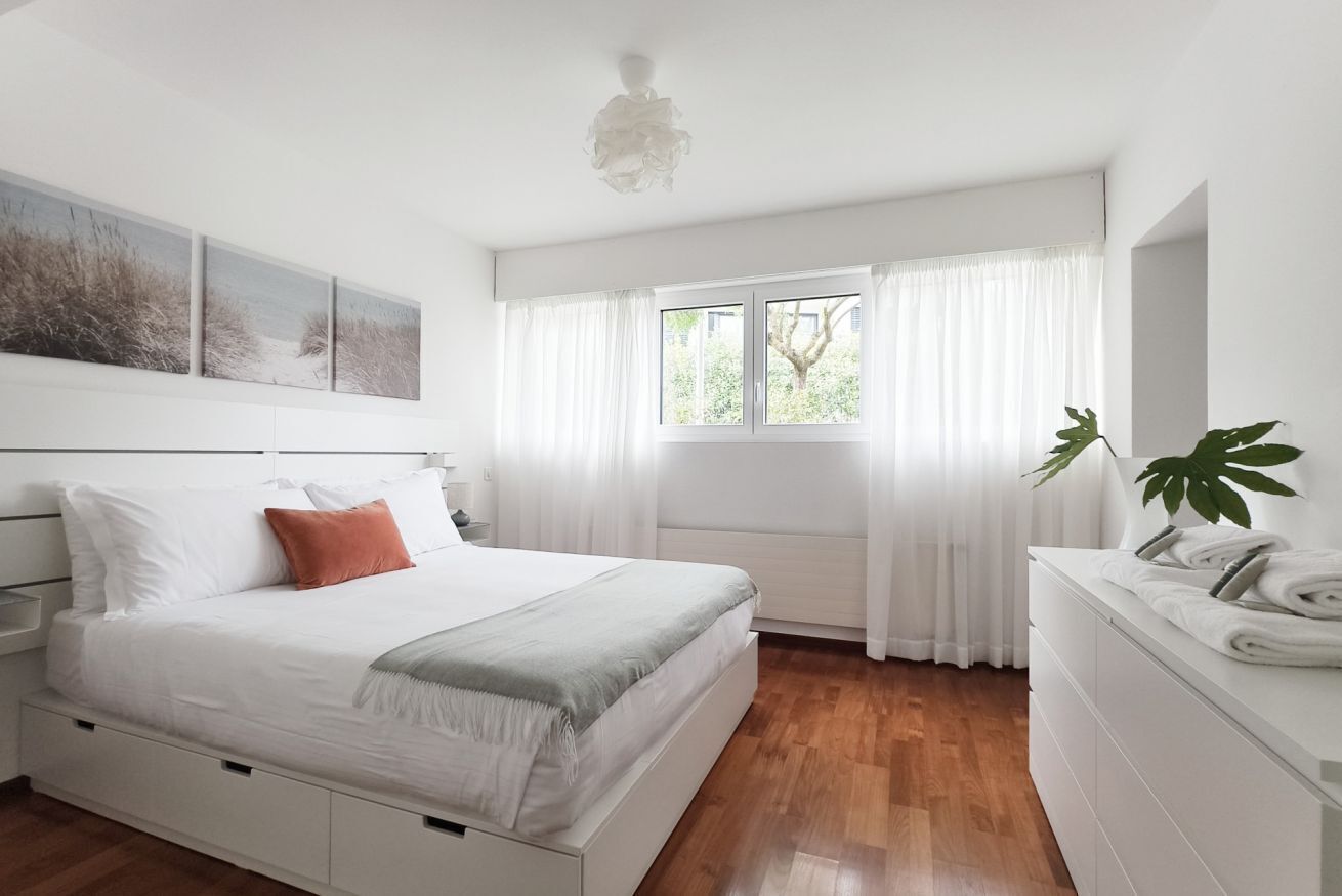 A bedroom with a bed, a nightstand, a dresser, a window with curtains, and a plant.