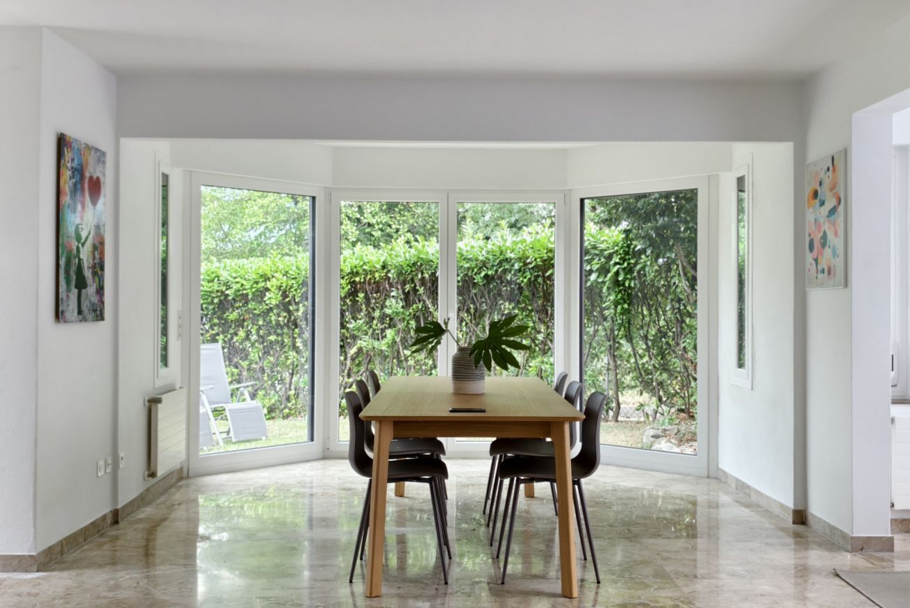 A dining area with a large table and chairs, large windows with a view of greenery, and artwork on the walls.