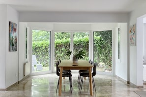 A dining area with a large table and chairs, large windows with a view of greenery, and artwork on the walls. - Apartment with garden near Lugano (Casa San Michele) (Muzzano)