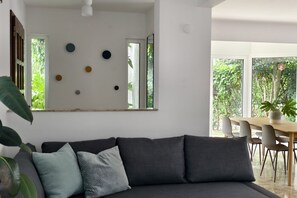 En moderne stue med en sofa, et spiseområde med stole og et stort vindue med udsigt til grøn vegetation.