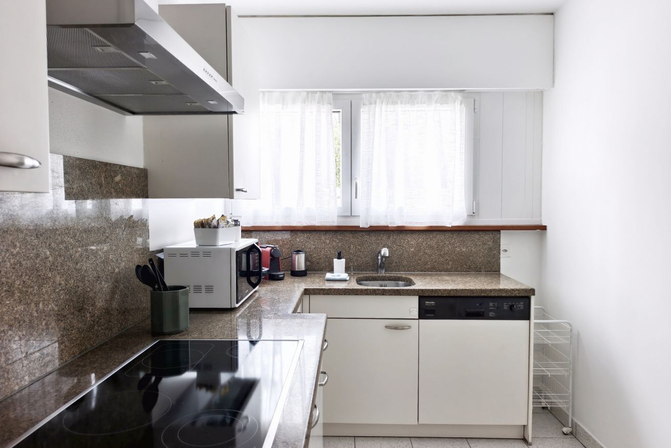 Ett modernt kök med bänkskivor i granit, en inbyggd diskmaskin och ett fönster med tunna, genomskinliga gardiner.