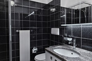 A modern bathroom with a black tiled wall, a white toilet, a sink with a countertop, and a shower area. - Apartment with garden near Lugano (Casa San Michele) (Muzzano)