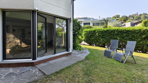Una casa moderna con amplios ventanales correderos, un camino de piedra y un césped cuidado con mobiliario exterior.
