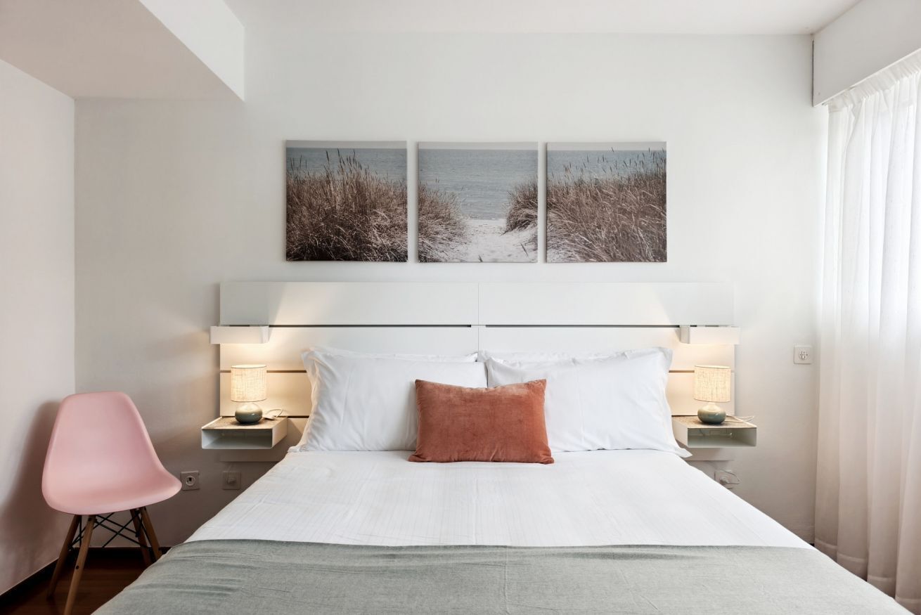 A bedroom with a bed, a nightstand, a lamp, a chair, and a beach-themed wall art.