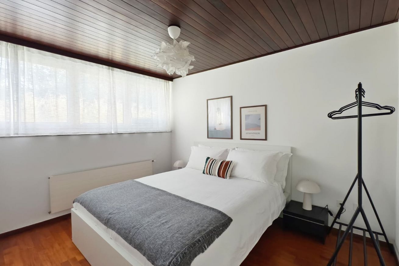 A bedroom with a bed, a radiator, a window with curtains, a hanging light fixture, and a coat rack.