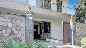 Un edificio moderno de dos pisos con fachada de piedra, entrada protegida por una reja y un pequeño jardín con plantas en macetas.