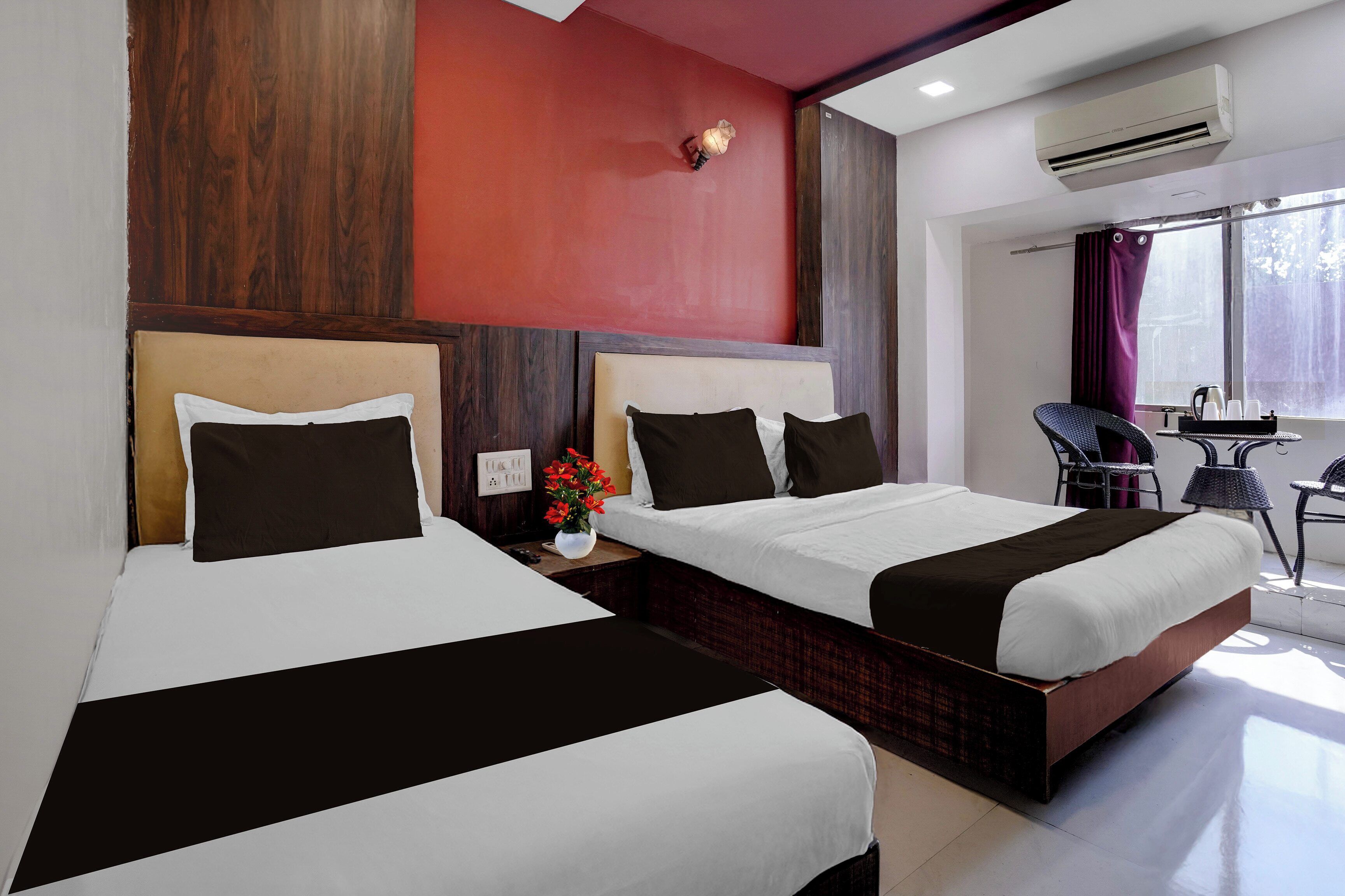 A hotel room with two beds, a red accent wall, an air conditioning unit, a window with curtains, and a small table with a vase of flowers.
