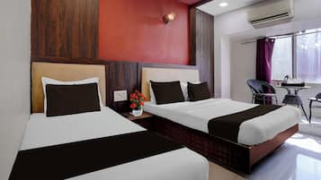A hotel room with two beds, a red accent wall, an air conditioning unit, a window with curtains, and a small table with a vase of flowers.
