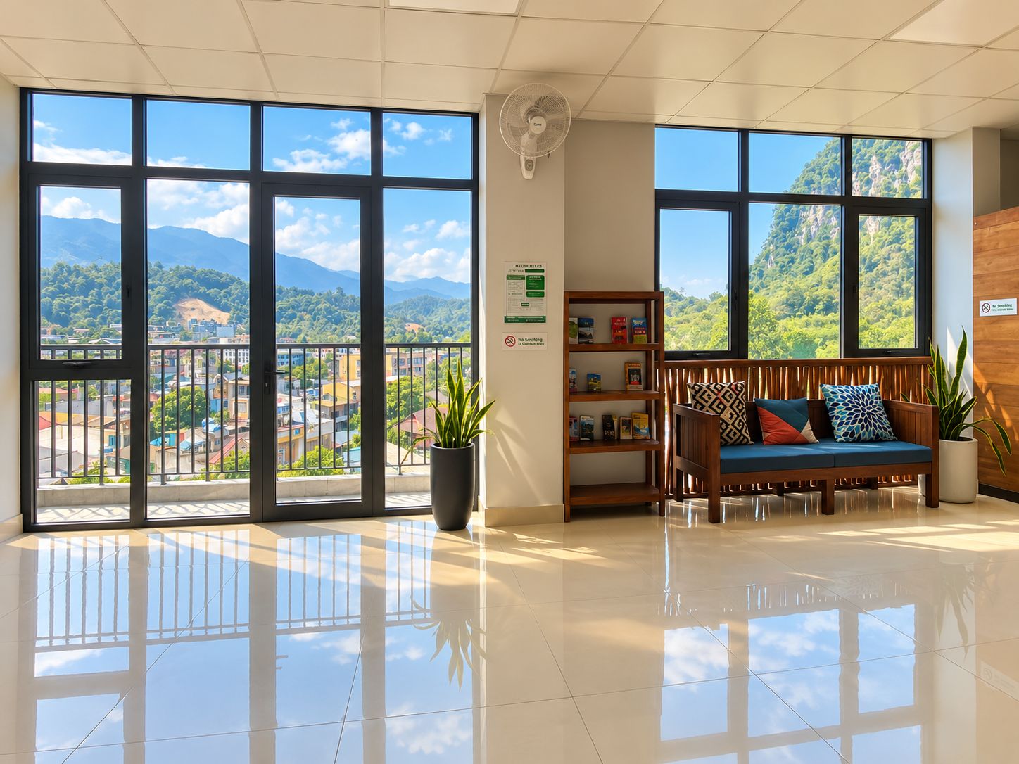 A hotel lobby with large windows, a reception desk, a seating area with a sofa and cushions, and a wall-mounted fan.