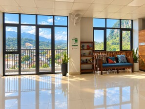 A hotel lobby with large windows, a reception desk, a seating area with a sofa and cushions, and a wall-mounted fan.
