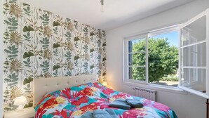 Un dormitorio con cabecera estampada con motivos florales, un ventanal con vistas a árboles, y una cama con edredón floral y toallas dobladas.