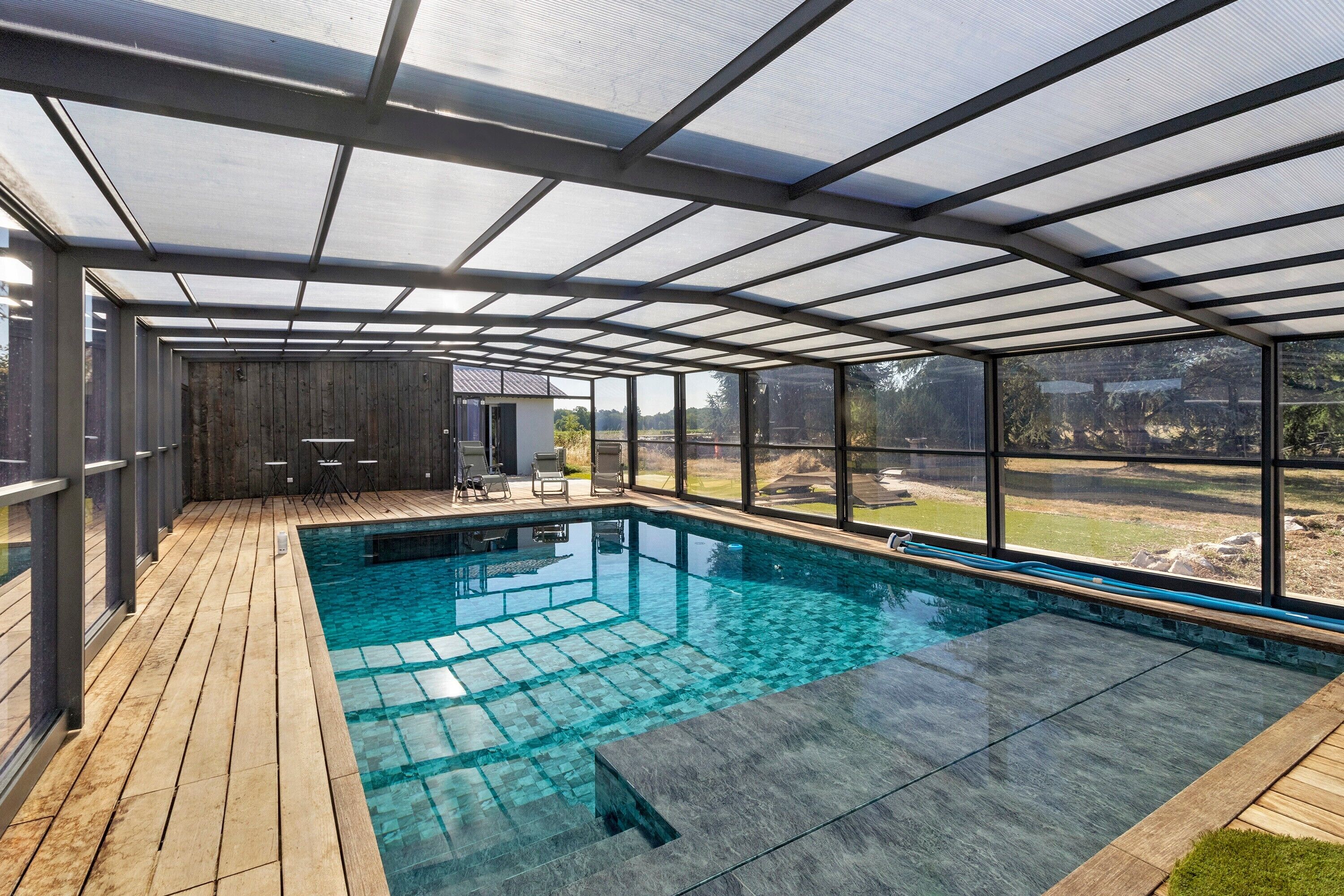 A covered swimming pool with a wooden deck, glass walls, and a view of the outdoors.