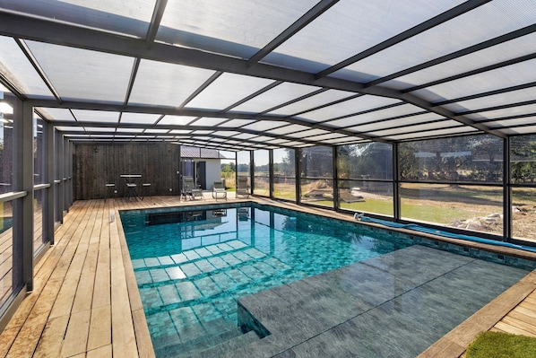 A covered swimming pool with a wooden deck, glass walls, and a view of the outdoors.