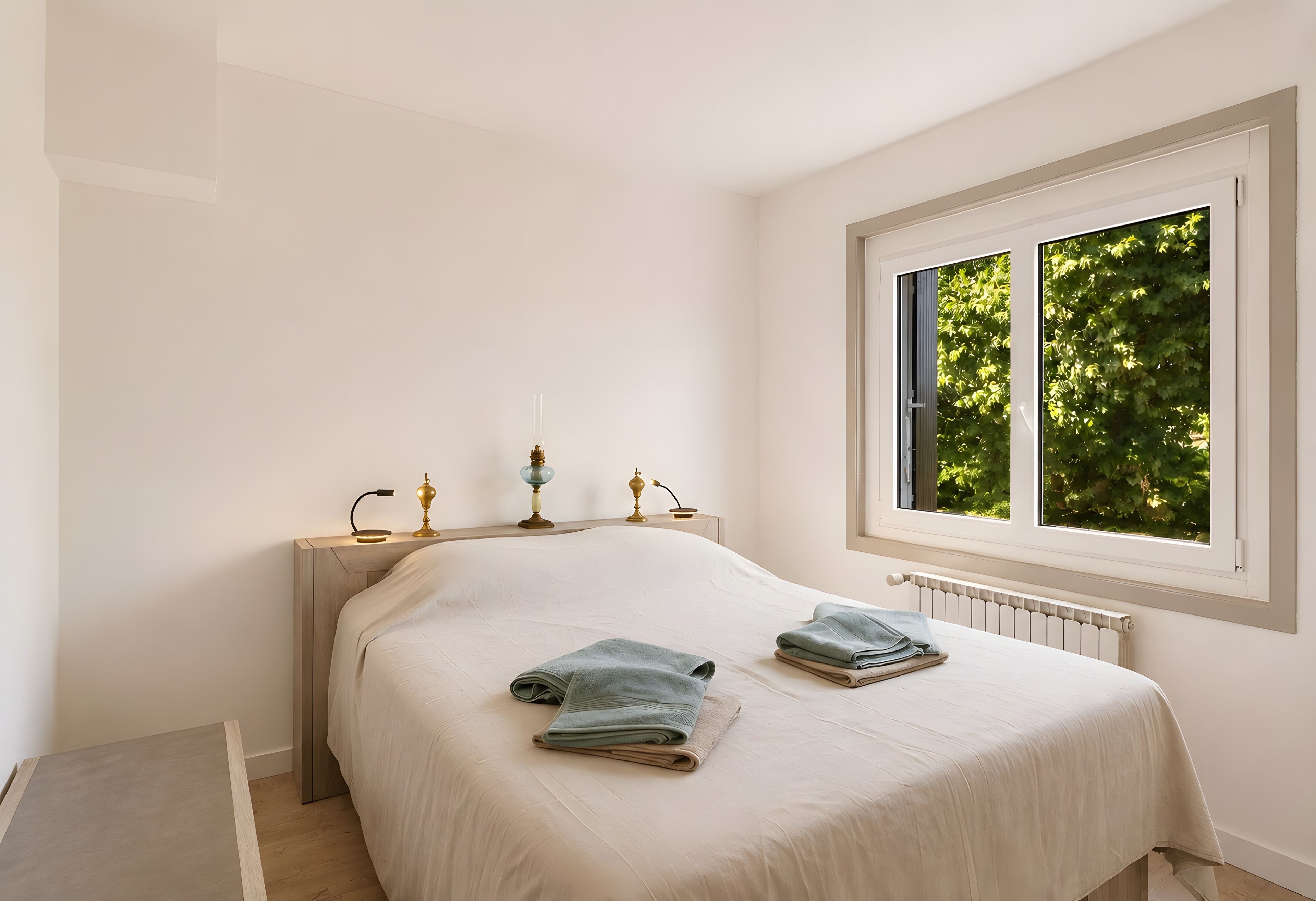 Een slaapkamer met een bed, twee bedlampen, een raam met uitzicht op bomen en een radiator.