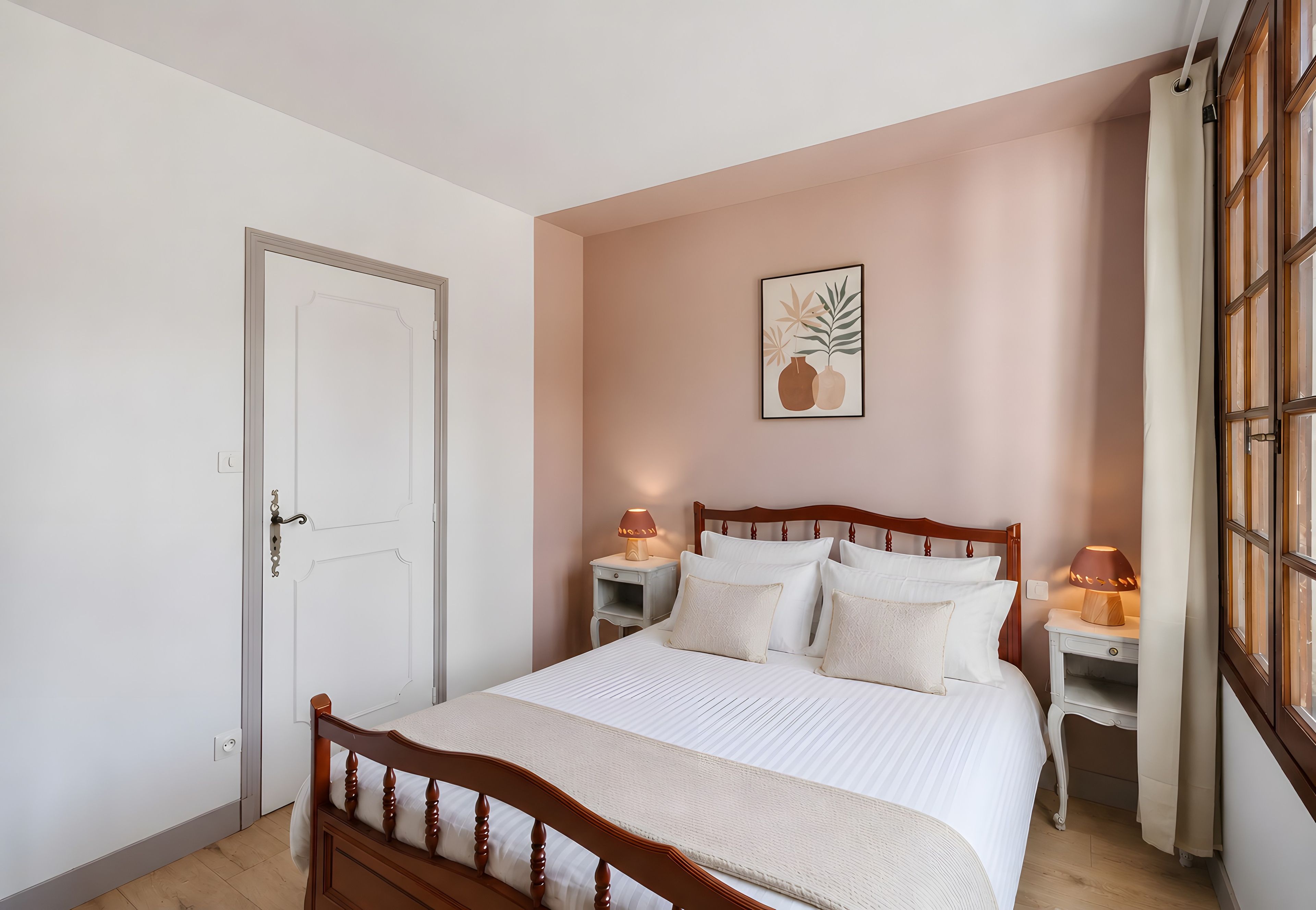 Een slaapkamer met een houten bed, witte beddengoed, twee nachtkastjes met lampen, een deur en een aan de muur bevestigd schilderij.