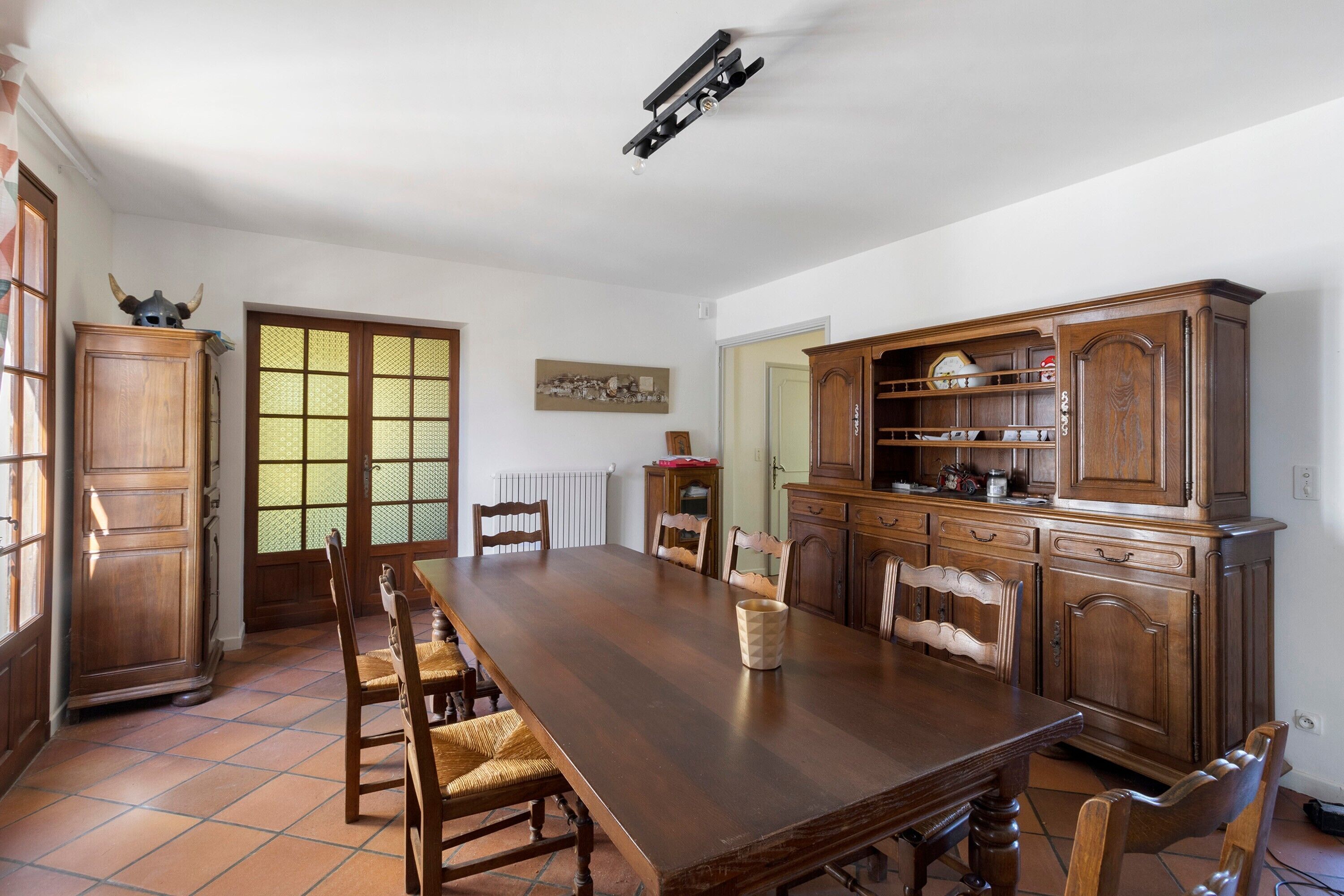Een eetkamer met een grote houten tafel, stoelen, een kast en een deur met een decoratief glasraam.