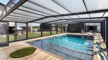 Una piscina techada con terraza de madera, paredes de vidrio y vista al jardín.