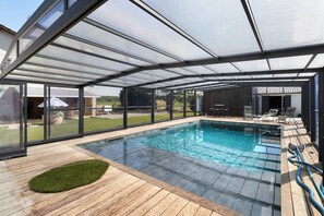 Una piscina techada con terraza de madera, paredes de vidrio y vista al jardín.