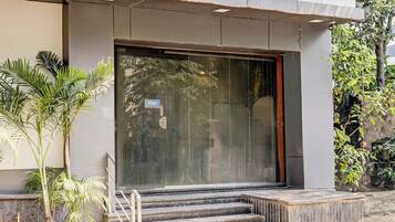 La entrada de un edificio con un amplio puerta corredera de vidrio, un conjunto de escalones que suben a ella y plantas en macetas a la izquierda.