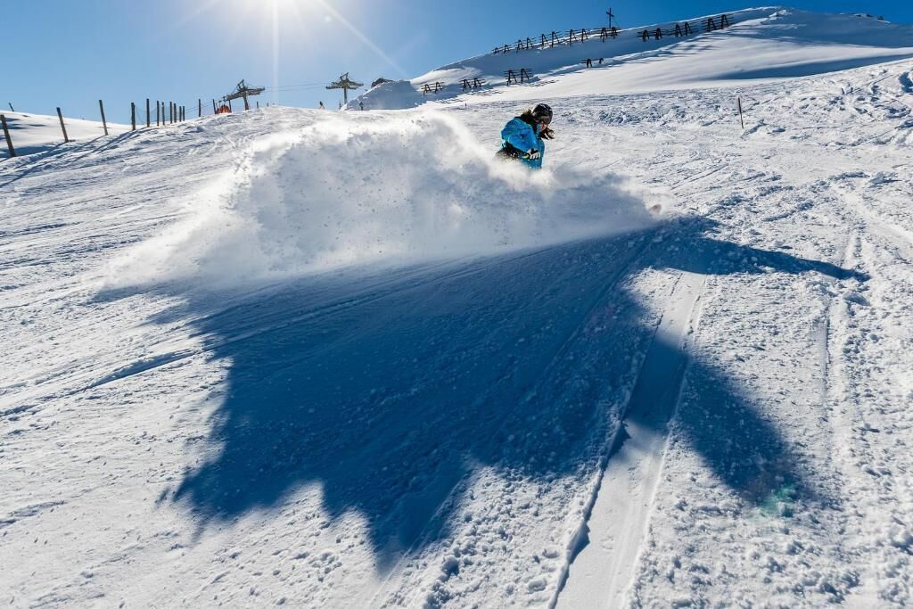 Un snowboardeur dévale une pente enneigée, le ciel est dégagé et une clôture est visible au loin.