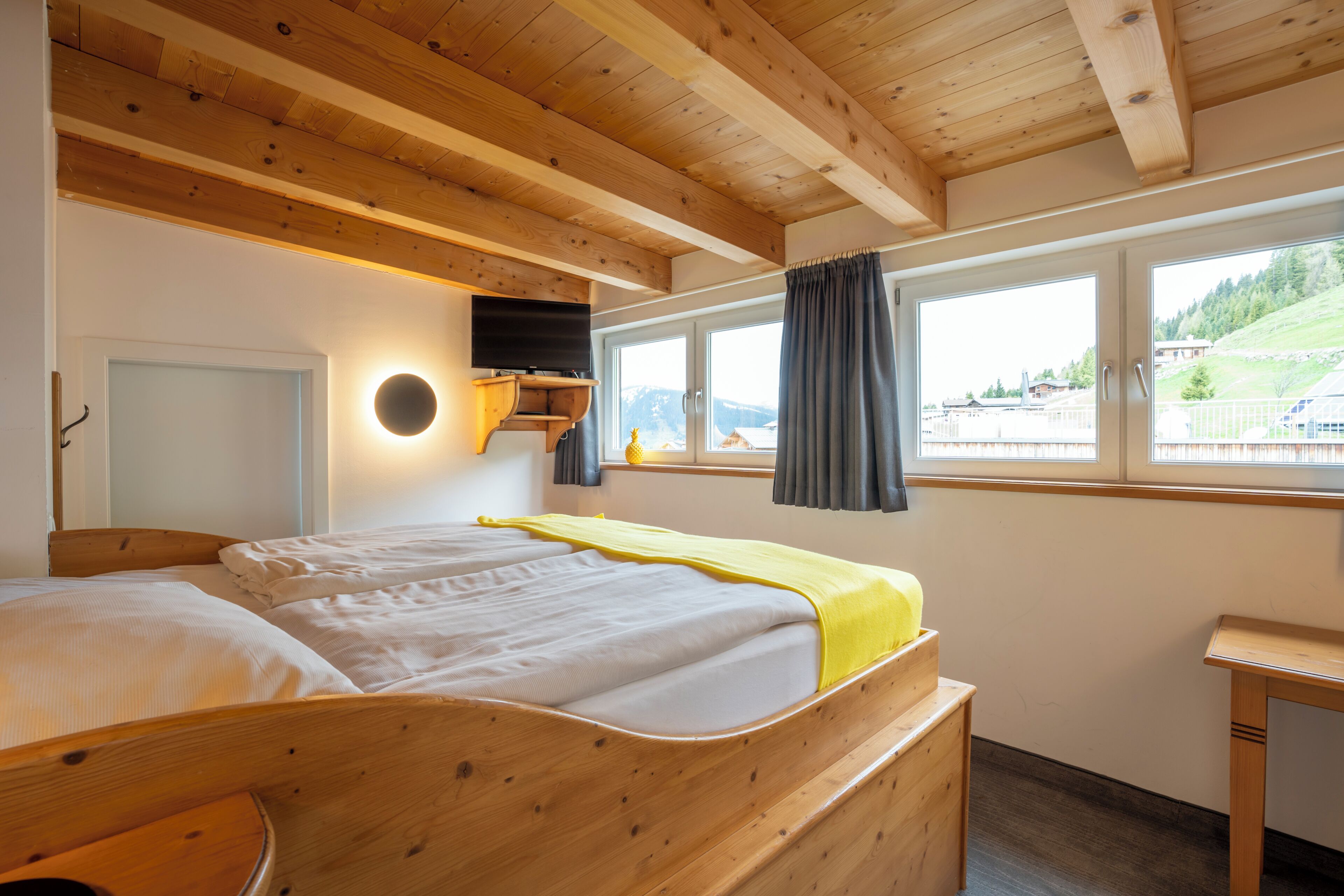 Ein Bett mit Holzkopfende, ein Nachttisch, ein an der Wand befestigtes Fernsehgerät und ein Fenster mit Vorhängen.