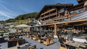 An outdoor seating area with wicker furniture and a bar, surrounded by a wooden building with balconies. - MY ALPENWELT Resort – 4 Sterne Superior (Königsleiten)